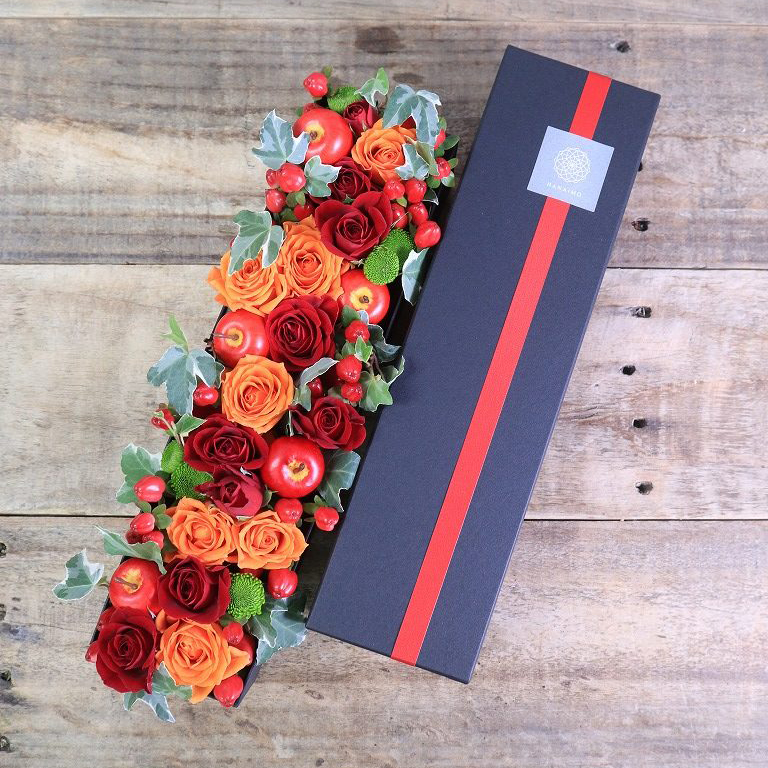 長方形のグレーの箱に赤やオレンジの花がぎゅっと詰め込まれたフラワーアレンジメント。右側には蓋がおいてあります。